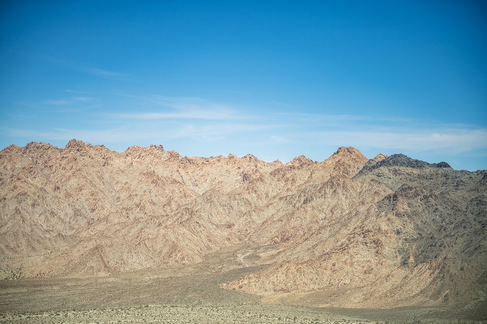 Bare tan desert mountains with dry washes at their foot, seen from the air under a blue sky