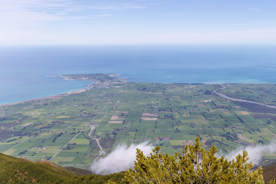 From a mountainside, a patchwork of green fields running to a turquoise sea, with a small peninsula and town jutting out at left