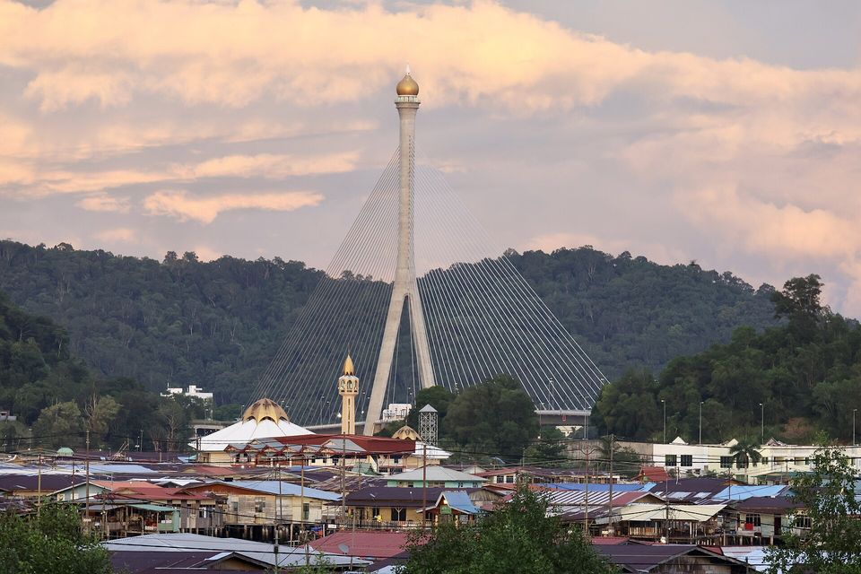 Rooftops of a water village crowd the foreground below a gold-domed mosque and minaret, a tall cable-stayed bridge pylon and forested hills under a pink evening sky