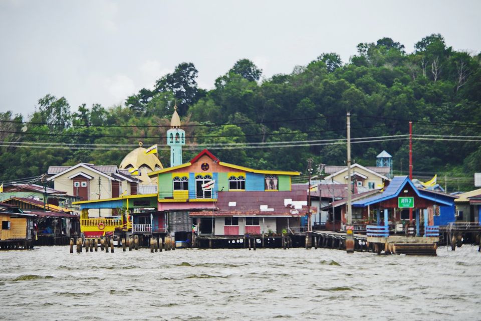 Brightly painted stilt houses in green, yellow, blue and pink cluster around a small mosque with a gold dome and a pale green minaret, forest behind, choppy brown water in front