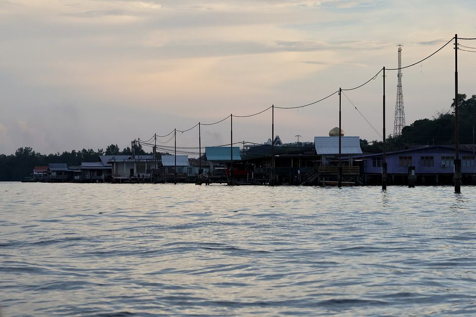 A row of dark stilt houses with pale roofs stands over silvery water at dusk, power lines strung on poles across the sky, a gold dome behind