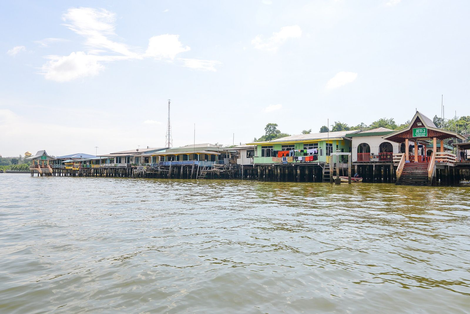 From a water taxi, a long row of pastel stilt houses on timber piles over the brown Brunei River, a roofed jetty stair at right, a pale tropical sky