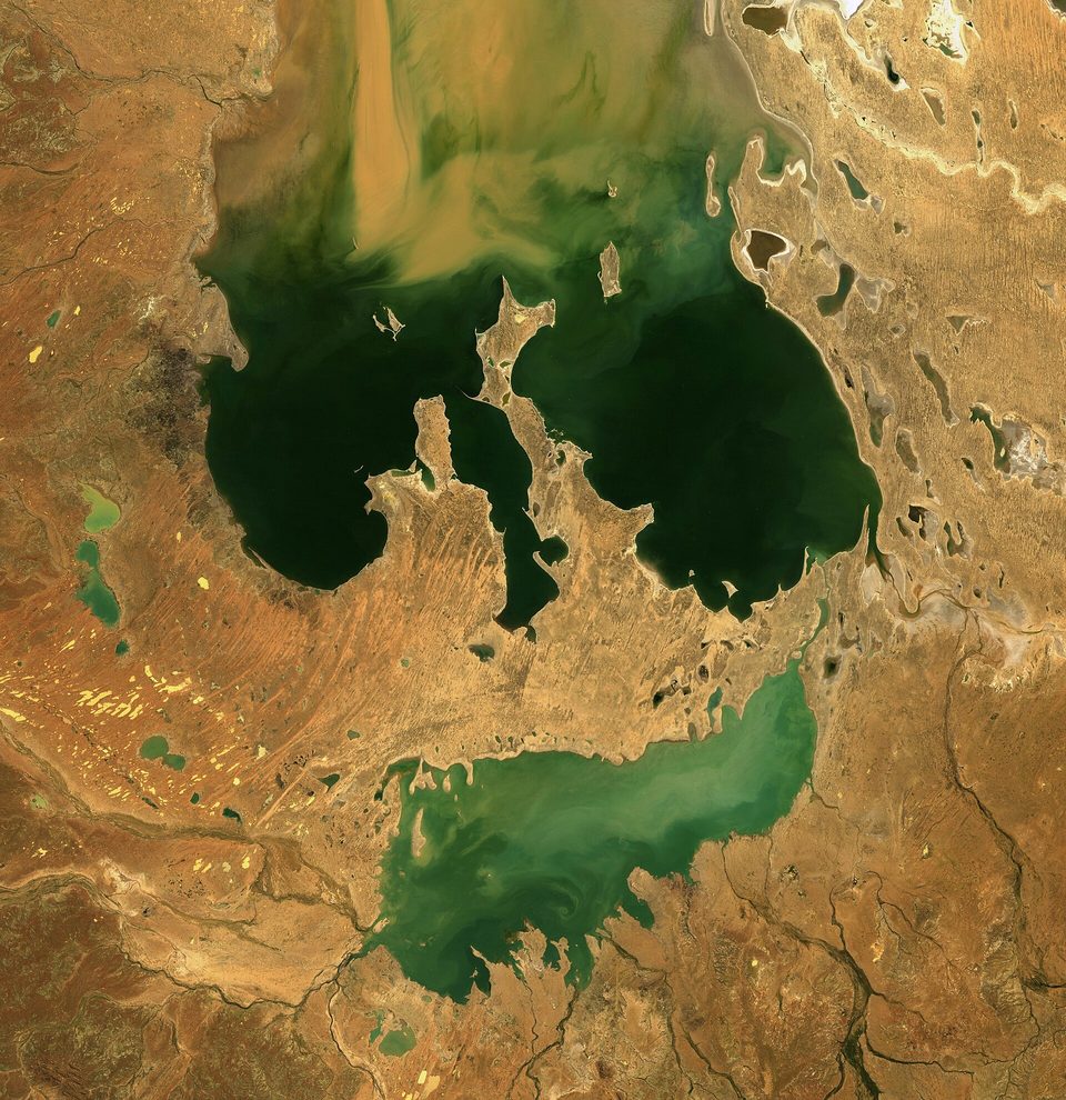 A true-colour satellite view of a large desert lake holding dark green floodwater, with pale shallows and red-orange desert around it