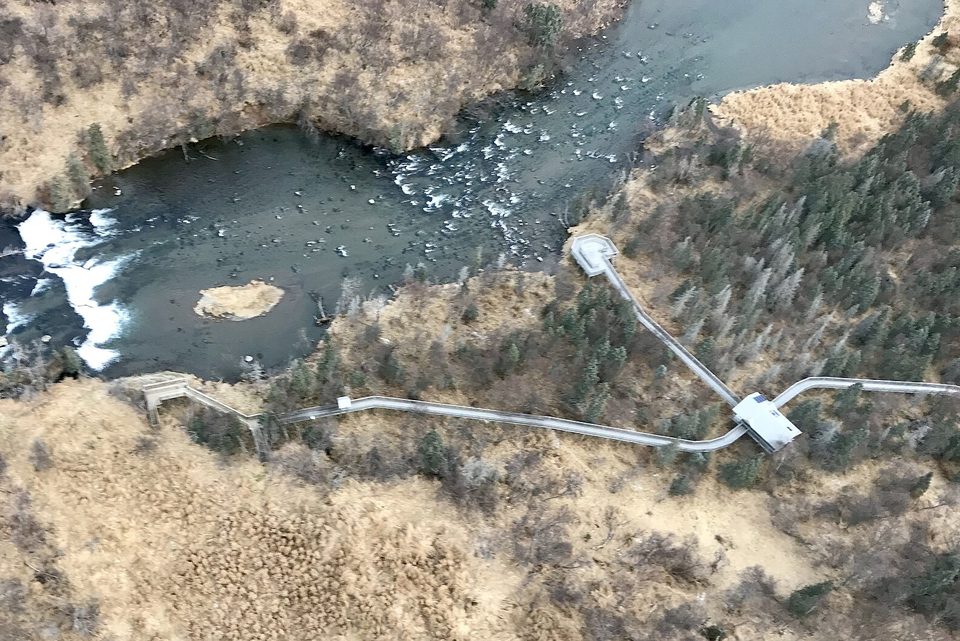 Looking down on a low wide waterfall in a dark river, a boardwalk and a raised platform running through autumn-brown brush beside it