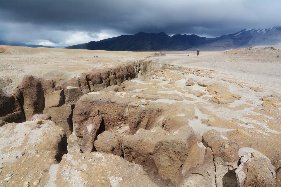 A wide plain of pale volcanic ash split by a deep gorge, a tiny hiker in the distance and dark mountains under storm cloud