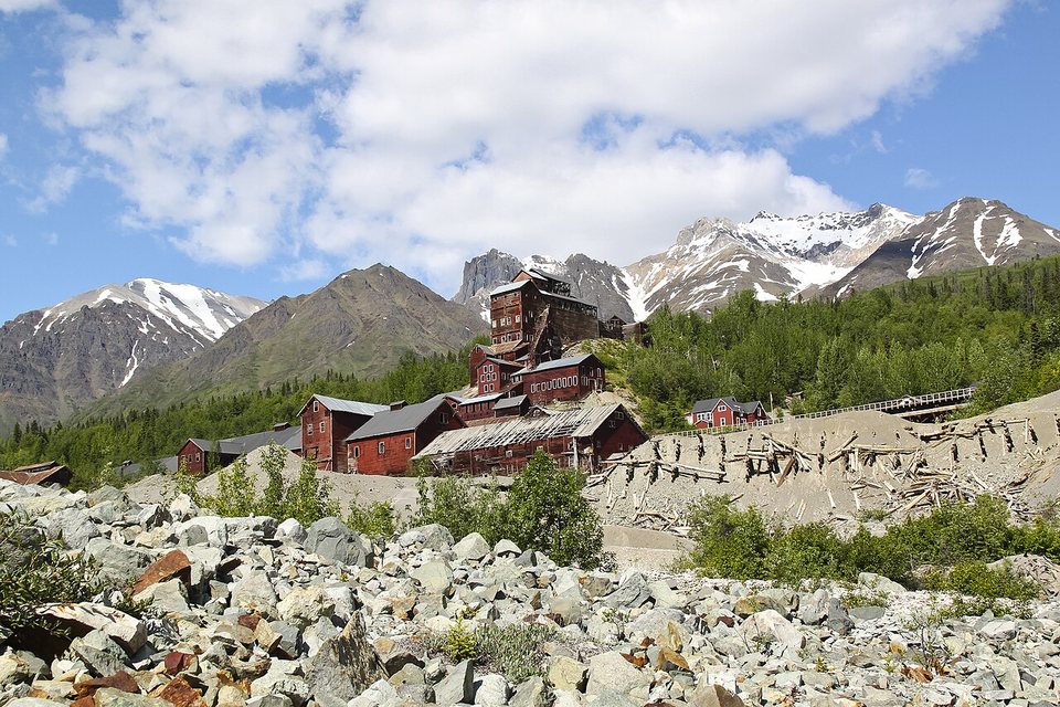 A red mill town below mountain peaks, seen across grey moraine boulders