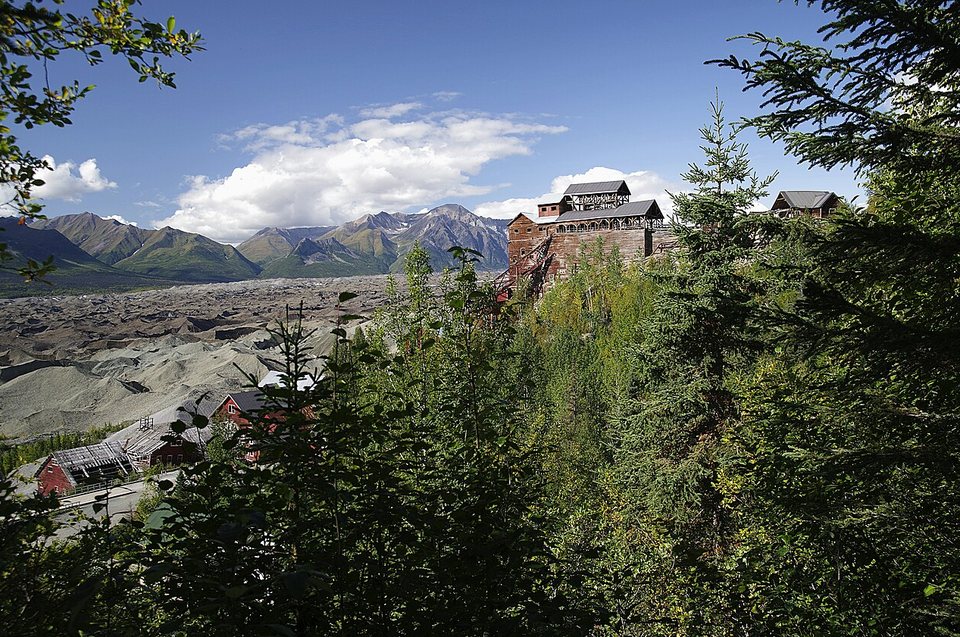 A tall red mill building framed by spruce, with a grey glacier and snowy peaks behind