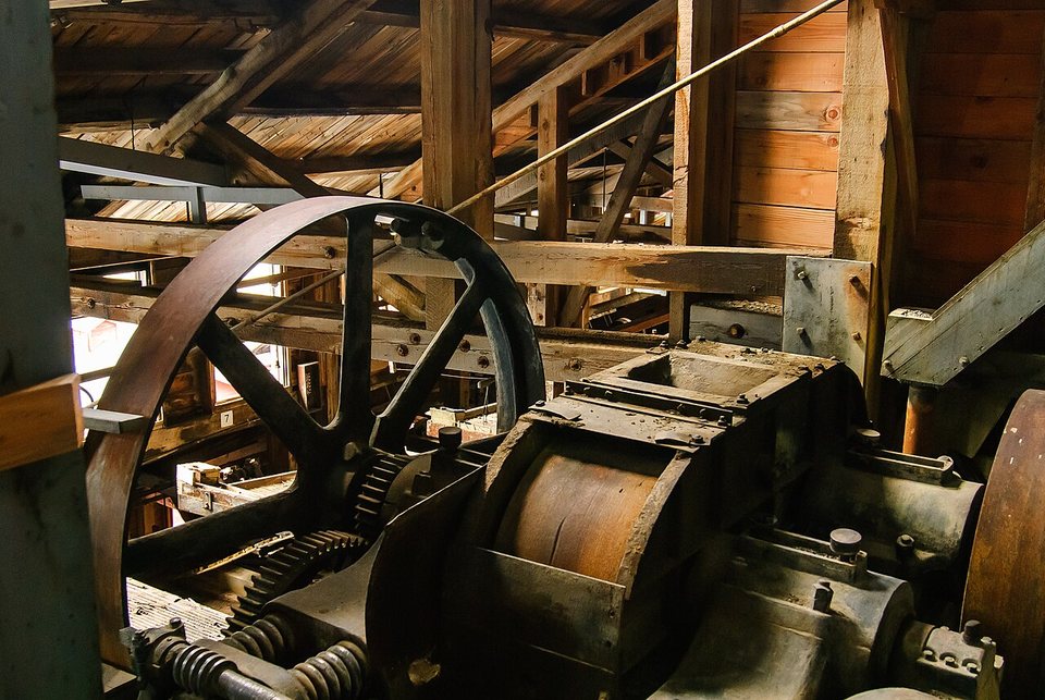Rusted iron machinery and a large wheel among timber beams inside a mill building
