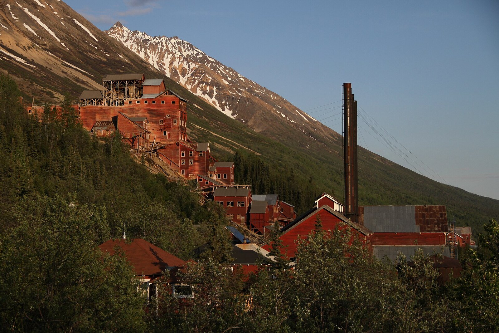 A red timber mill and town stepping down a slope in warm low light, with a snow-streaked peak behind