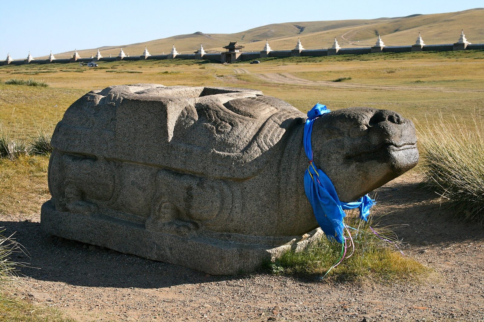 A granite turtle with a blue ceremonial scarf at its neck in short steppe grass, a white stupa wall behind and low hills under a pale sky