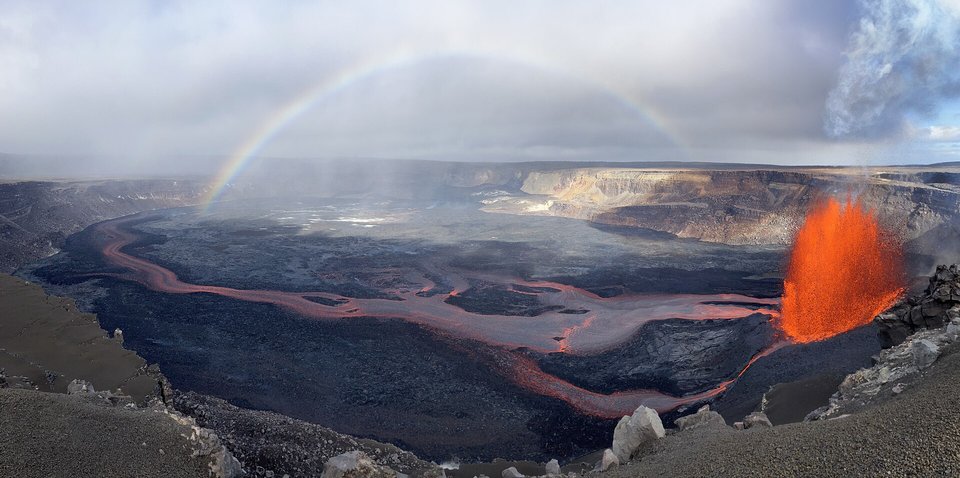 A rainbow arching over a wide dark crater floor crossed by glowing lava channels, a fountain of orange lava at the right and a pale cloud at the top