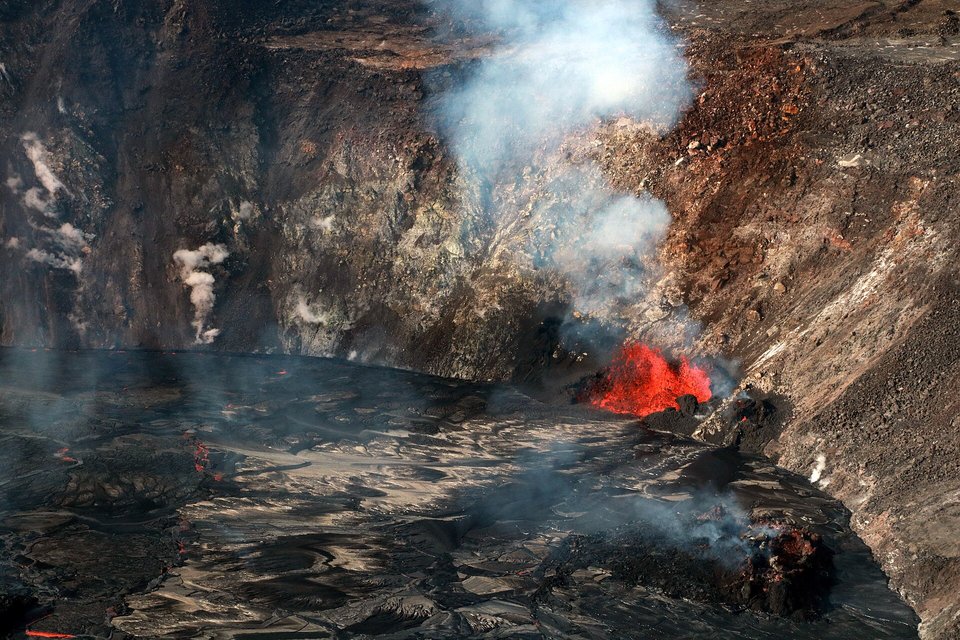 An aerial view into a steep-walled crater with a dark crust of lava lake on its floor, a glowing red vent at the foot of the far wall and plumes of white gas rising