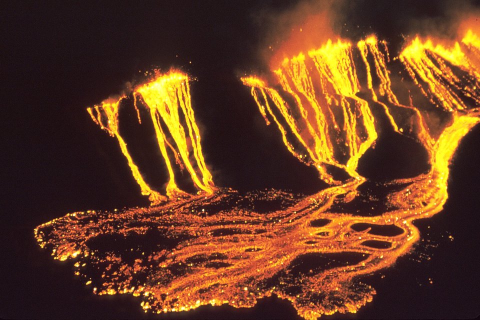 Glowing yellow and orange lava pouring in streams down a dark crater wall and spreading in a bright web across the floor at night