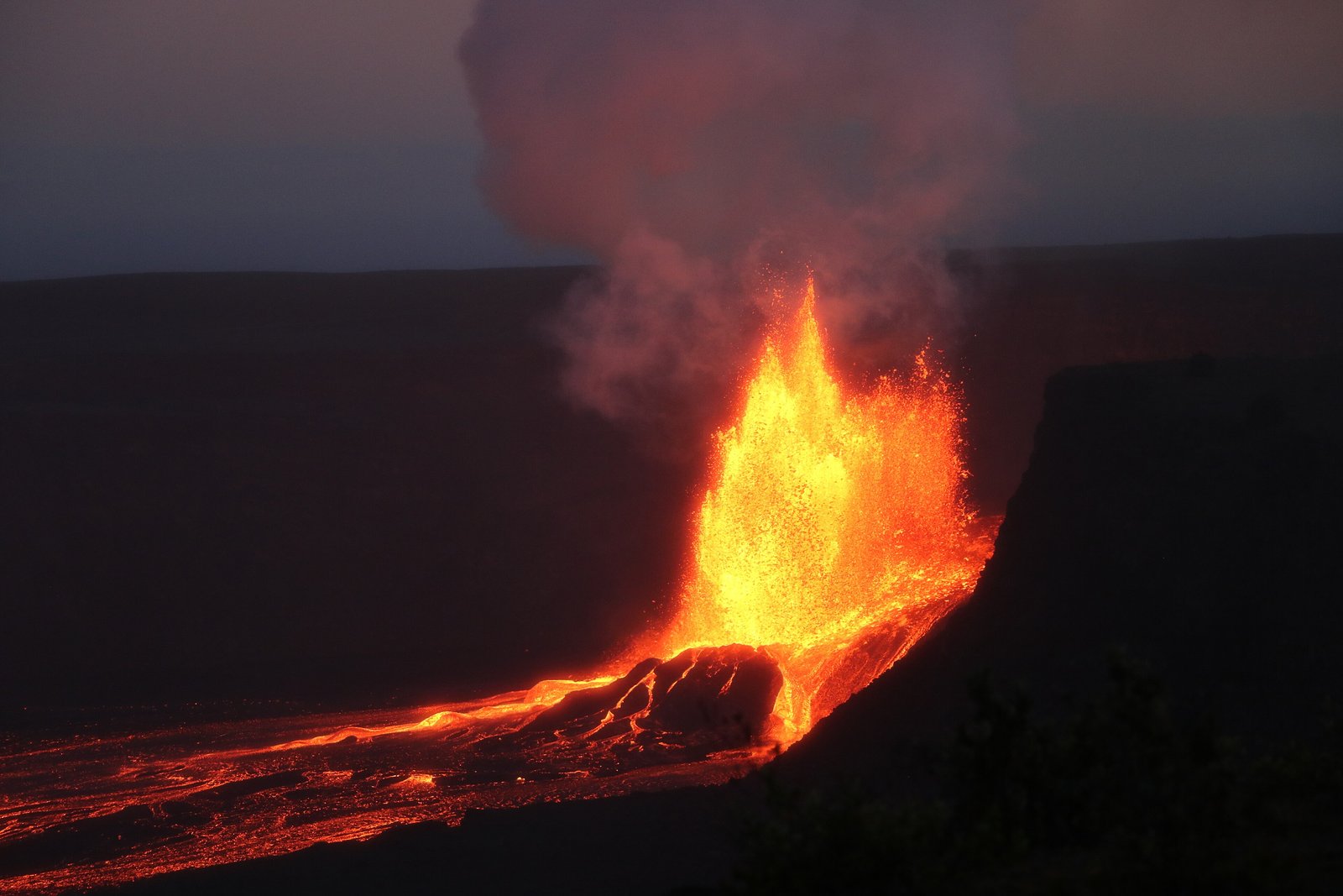 A tall fountain of glowing orange lava rising from a vent beside a crater wall, lava flowing across the dark crater floor and a column of smoke above