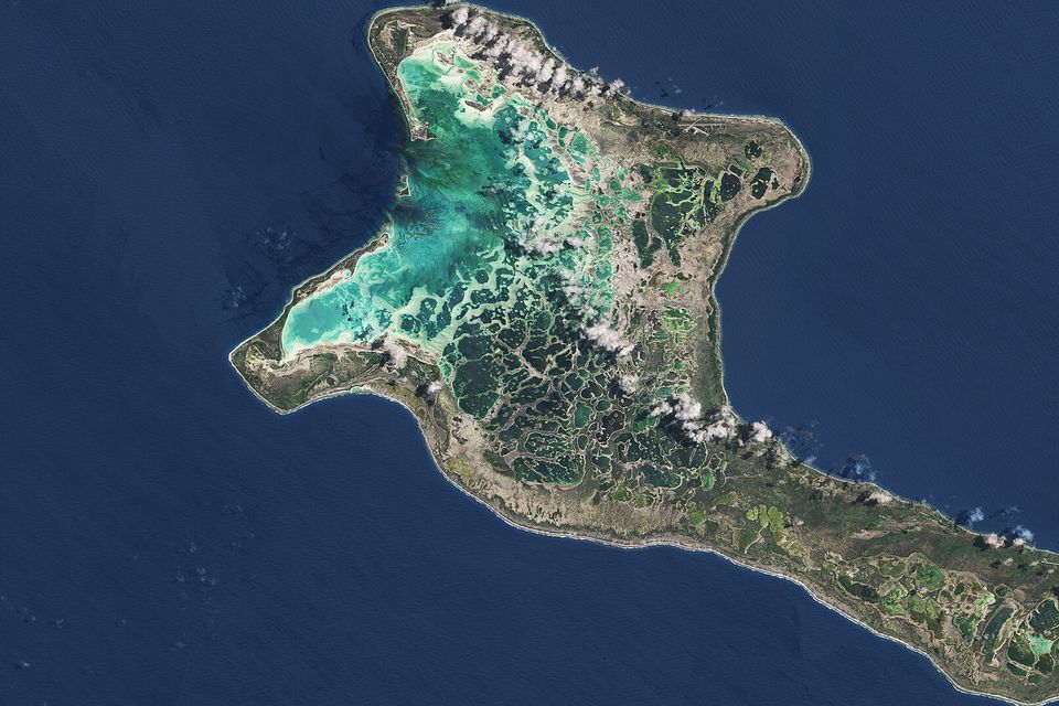 A hook-shaped atoll in dark blue ocean, its west side a bright turquoise lagoon and its interior a mosaic of green and brown ponds