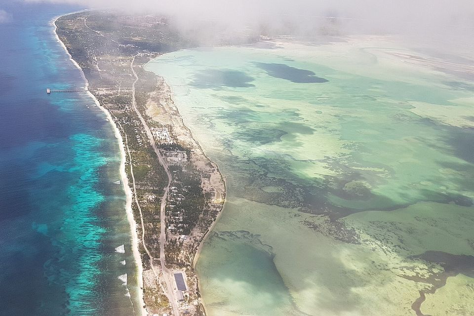 From the air, a narrow strip of land with a road and scattered roofs separates deep-blue surf on the left from a pale green lagoon on the right