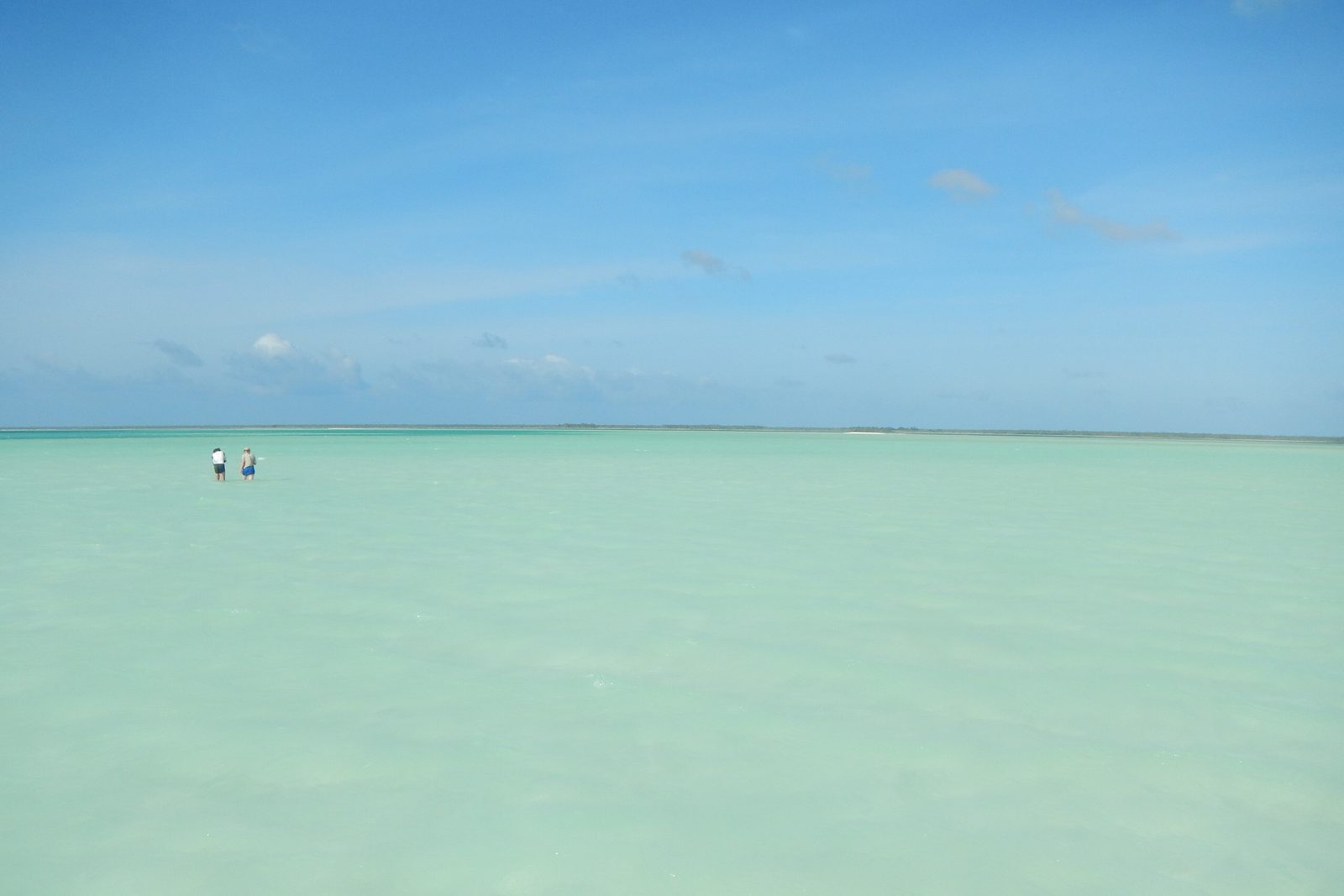 A sheet of pale jade lagoon water runs flat to a thin line of scrub on the horizon under a soft blue sky, two tiny waders far out at left