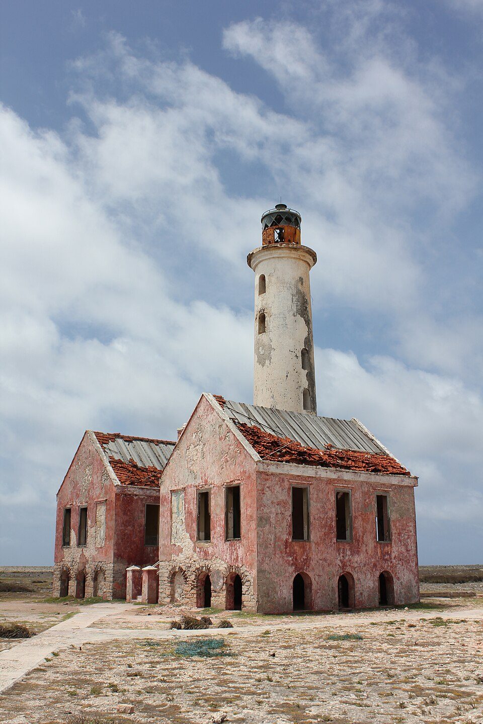 A tall white lighthouse rises from a two-gabled house of peeling pink plaster with a collapsing tiled roof, under broken cloud
