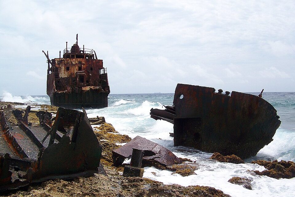 The rusted stern of a ship stands in breaking surf, with a broken bow section on the reef in front