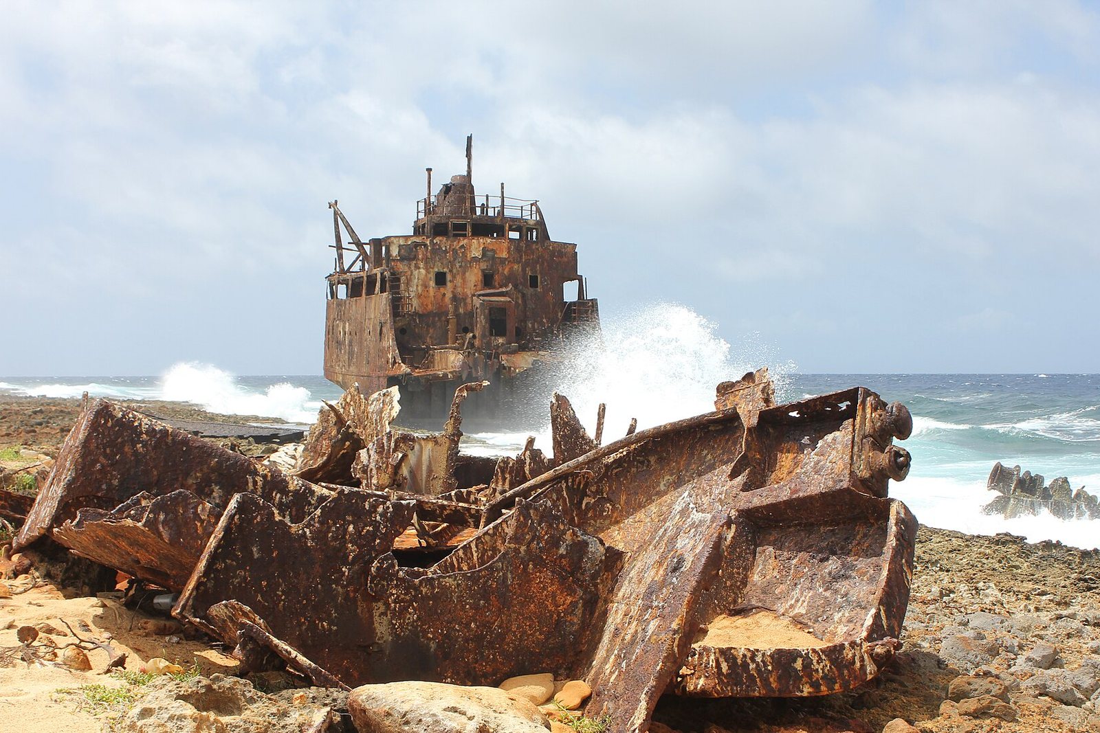 The rusted bow section of a stranded ship sits in the surf with spray bursting behind it, and torn hull plates lie on the coral shore in front