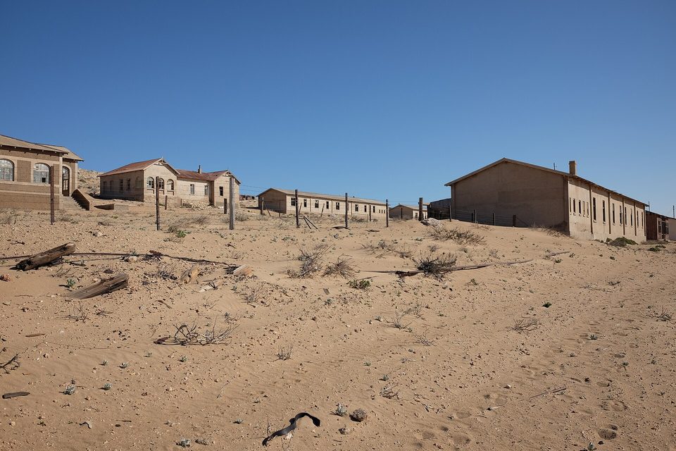Gabled stone houses in a row along a sandy ridge under a hard blue sky, fence posts and driftwood lying in the sand