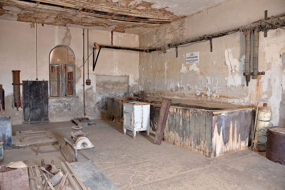 A plaster-walled industrial room with pipes along the ceiling and walls, a wooden counter, an arched window and old equipment on a dusty floor