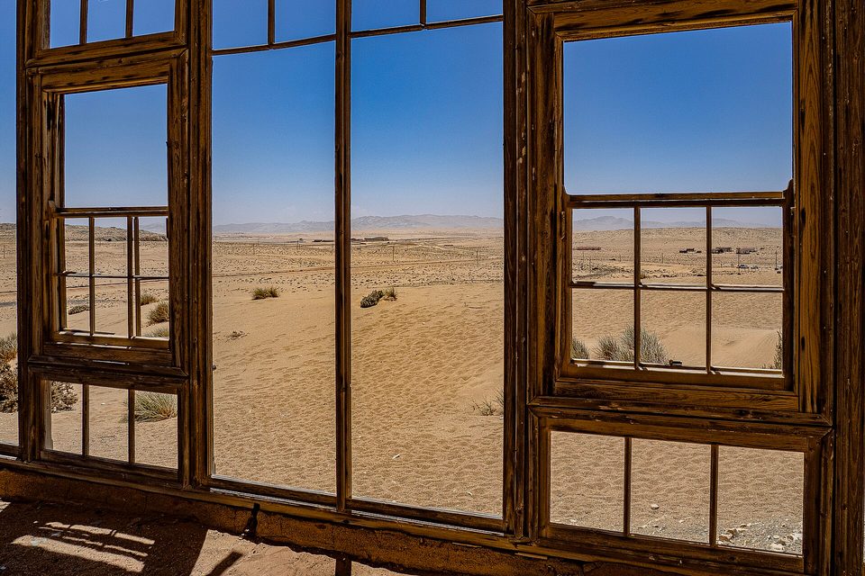 Wooden window frames with no glass looking out on flat sand and a line of far hills under a blue sky
