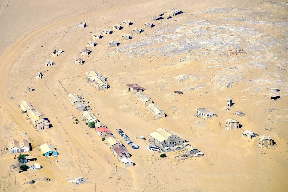 A scatter of small houses and a few larger buildings on pale sand, seen from above, with tracks curving through the settlement
