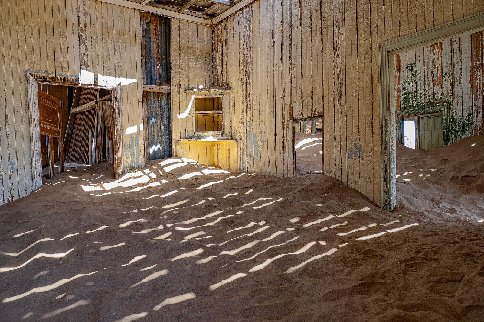 Pale sand drifted across the floor of a plank-walled room and through two open doorways, striped with sunlight from the broken roof