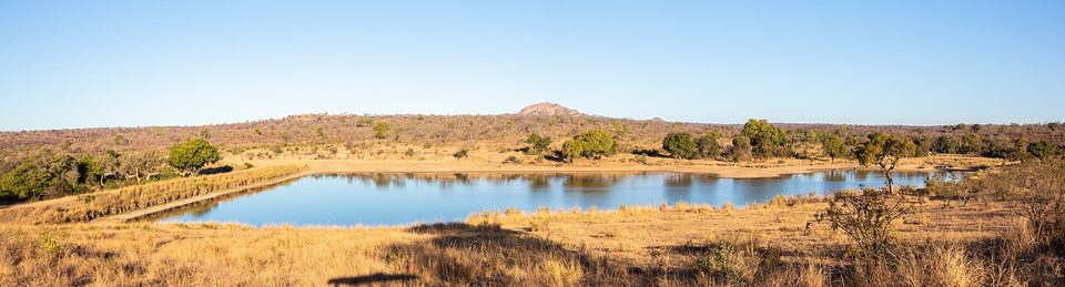 A wide dry-season landscape of golden grass around a blue waterhole with a low granite dome on the horizon under a clear sky