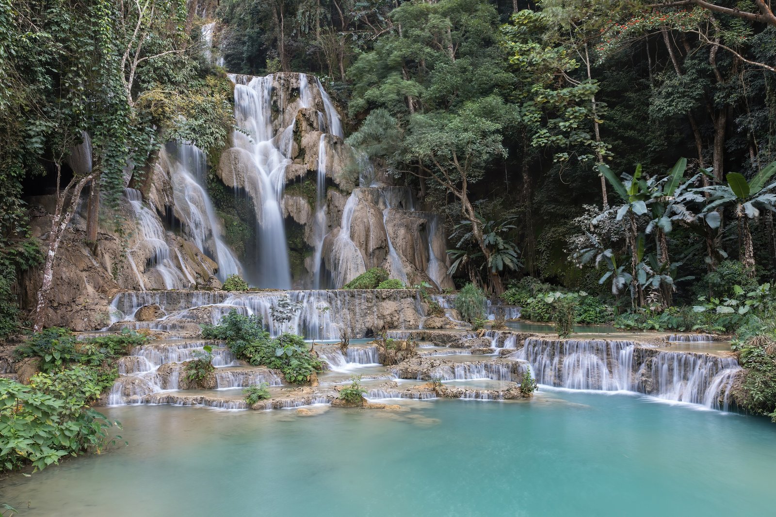 A tall waterfall stepping down pale limestone into a milky turquoise pool, with forest and banana leaves around it and low tiered cascades in front