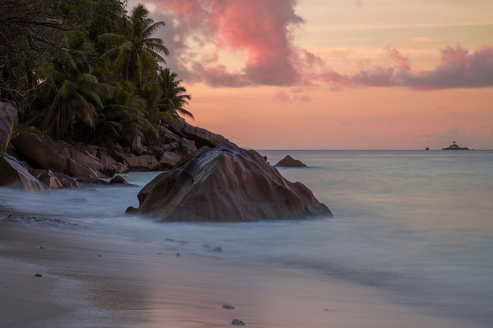 A rounded granite boulder in silky surf under a pink and orange evening sky, palms on the shore at left