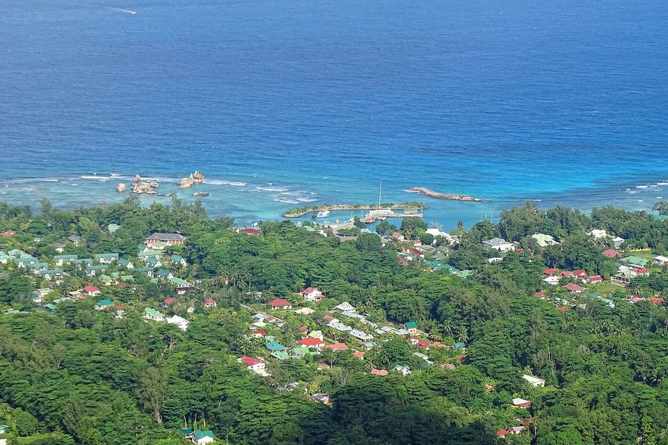A small harbour with a breakwater and boats on bright blue water, houses with red and green roofs among trees