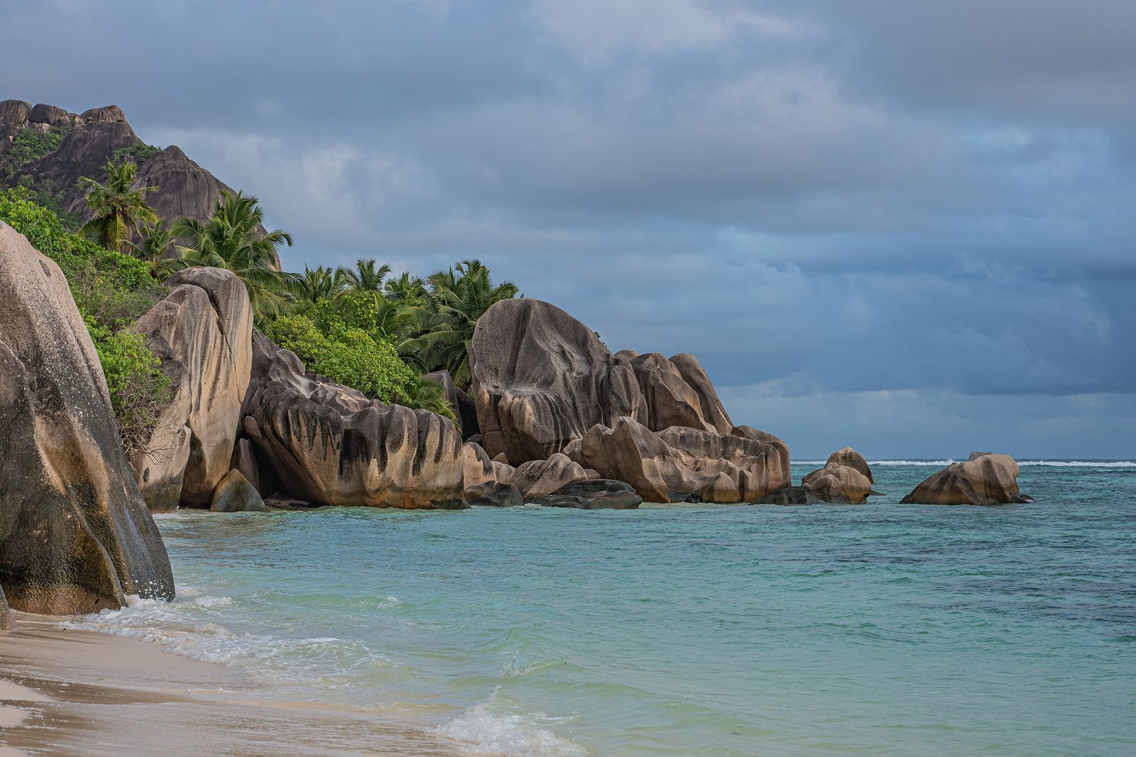 Smooth granite boulders along a beach with palms on the slope behind and turquoise water under a stormy sky