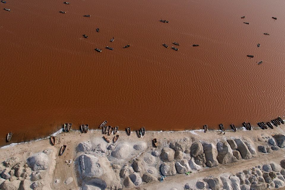 Red-brown water dotted with small wooden boats above a shoreline of grey and white salt heaps