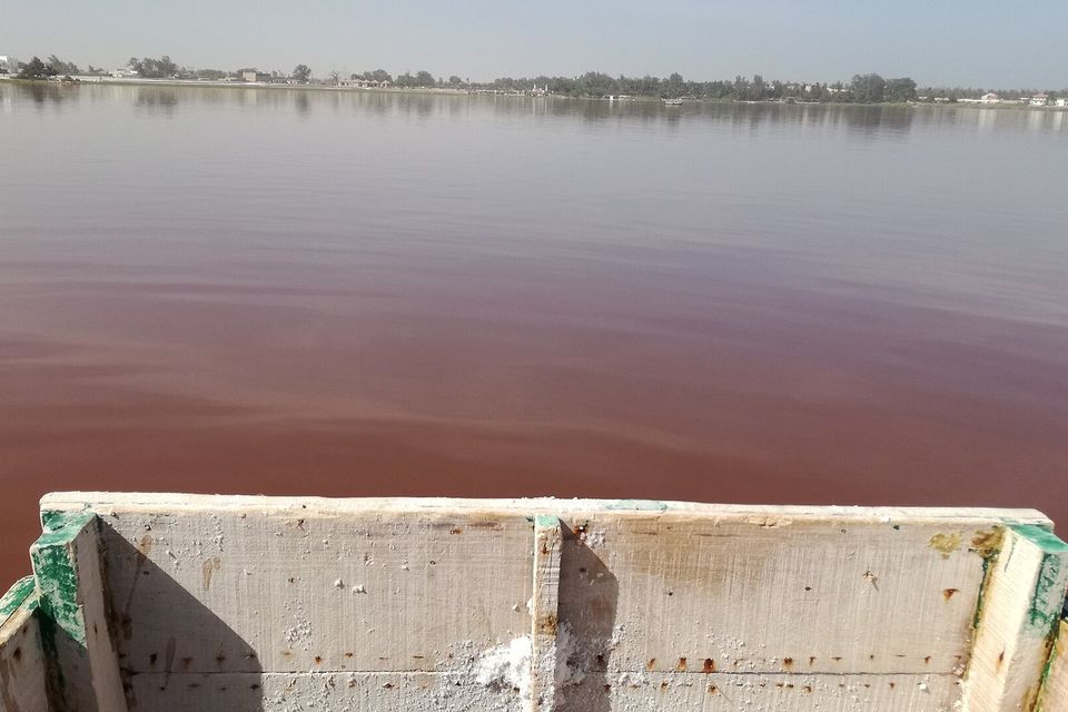 The salt-crusted planks of a boat bow in the foreground and a wide sheet of dusky red-brown water reaching a low tree-lined shore