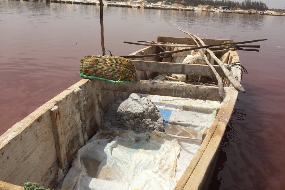 The inside of a wooden boat holding a heap of grey wet salt on a white plastic sheet, a woven basket across the gunwale, red-brown water around it