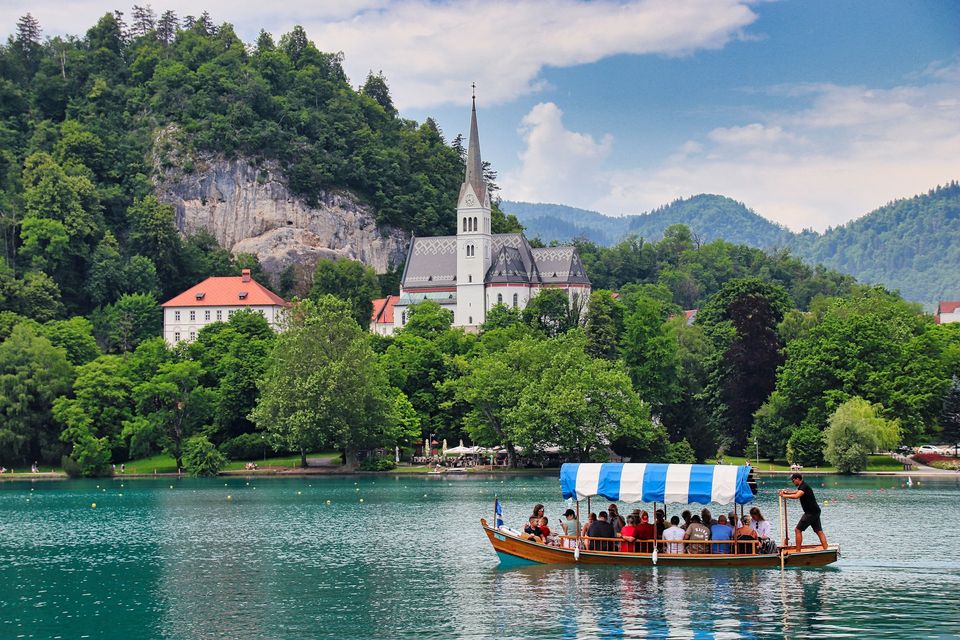 A canopied wooden boat with passengers and a standing oarsman on a lake beside a shoreline church