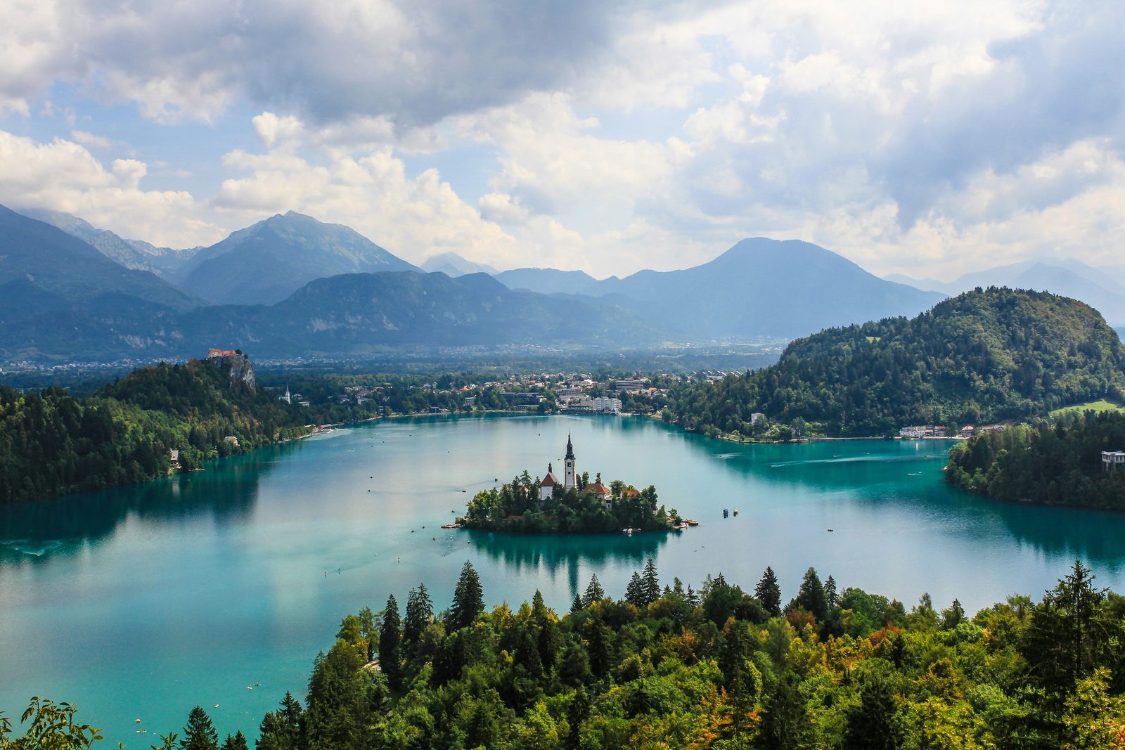 A turquoise lake with a small island holding a church, a castle on a cliff at the left and mountains behind under broken cloud