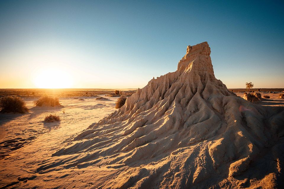 A tall cone of fluted, sun-lit sand and clay rises beside a flat desert plain, the sun low on the horizon