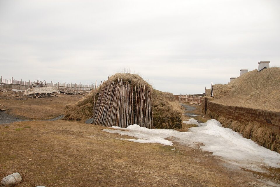 A low turf-covered structure walled with upright poles, beside patches of snow and a turf hall with chimneys, under a grey sky