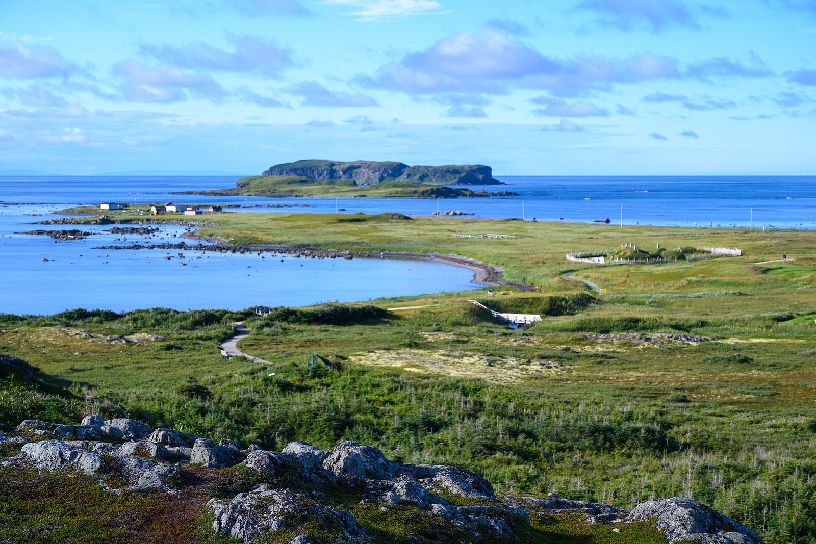 A granite outcrop and green barrens with a boardwalk above a blue bay, with the village and an island beyond