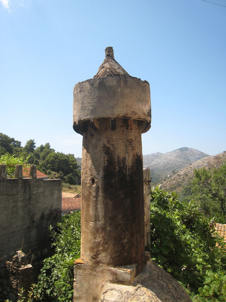 A tall round stone chimney with a crenellated cap and a pointed top stands against a clear blue sky, green hills behind