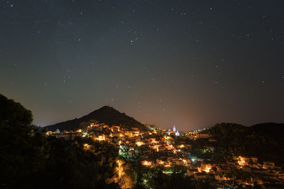 The lights of a hillside village glow amber below a dark sky full of stars, a pointed hill and a church tower in silhouette
