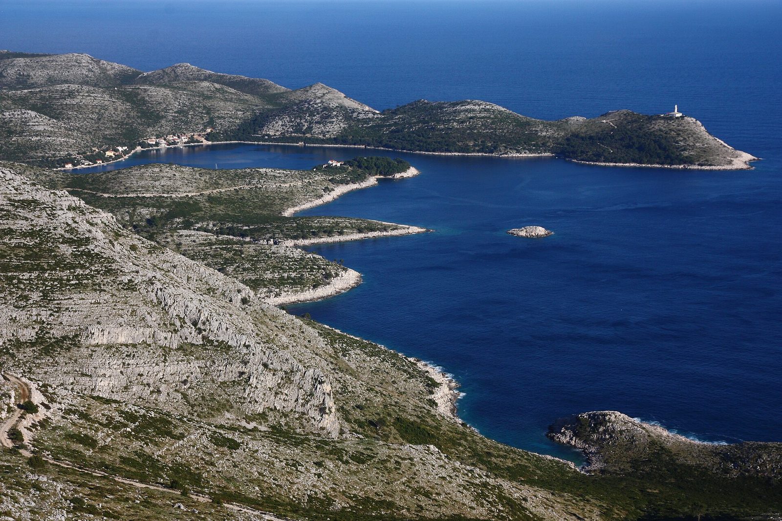 From a high limestone ridge, bare grey-green slopes fall to a sheltered bay of deep Adriatic blue, a tiny hamlet at its head, and a white lighthouse on the far headland above the open sea