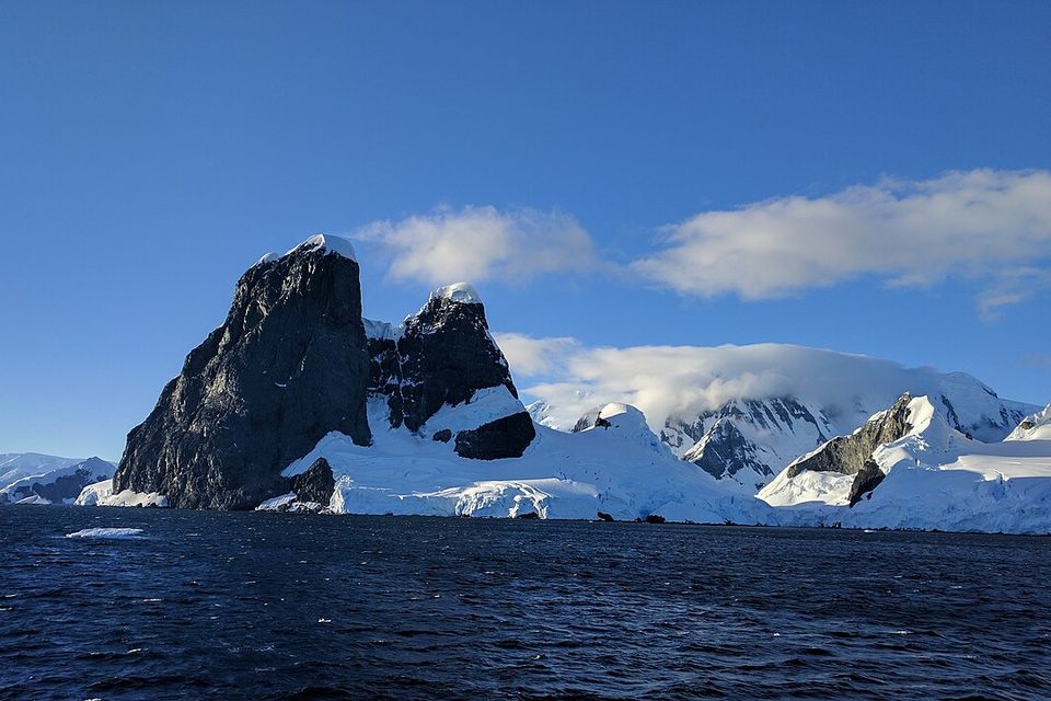 Dark, snow-streaked rock peaks rise above still water under a blue sky