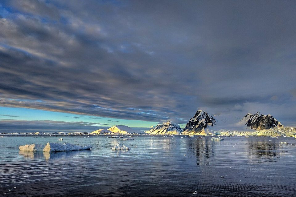 Low golden light catches snowy peaks across still water under a heavy band of cloud