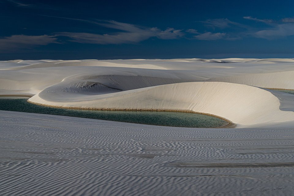 A pool of blue-green water lying in a hollow between pale rippled dunes under a deep blue sky