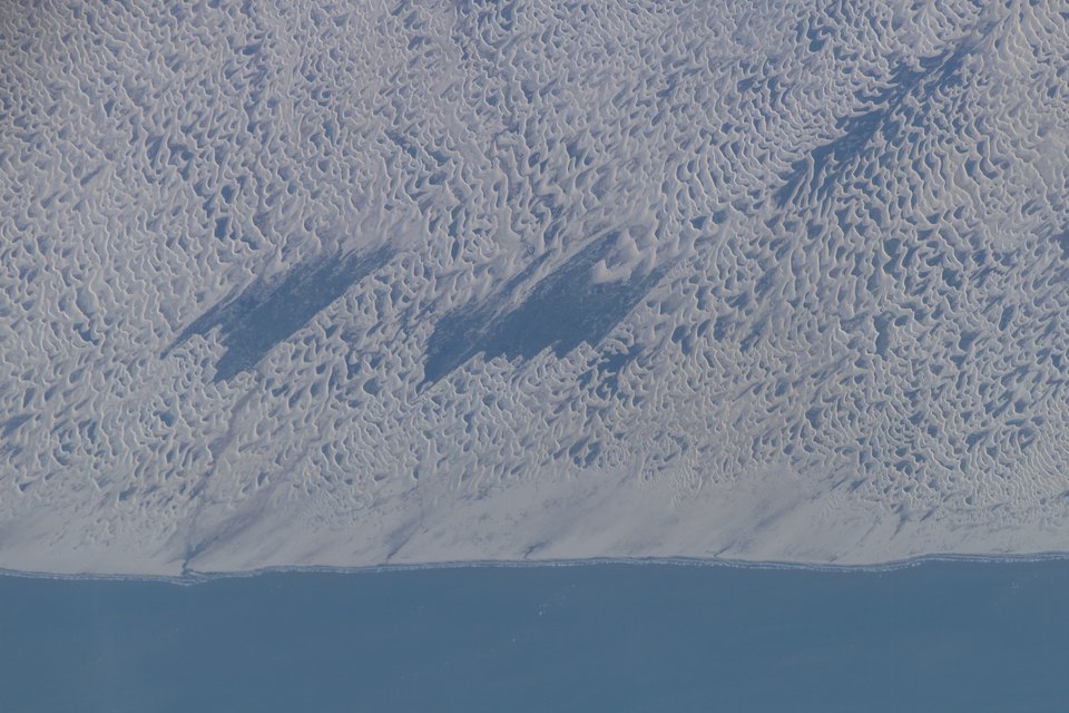 Pale rippled sand dunes running down to a straight blue shoreline, with dark cloud shadows lying across the sand