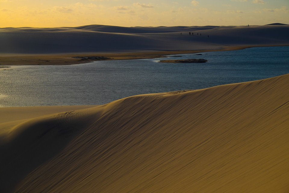 Pale dunes glowing in low evening light around a dark blue lagoon, with a line of tiny figures on the far ridge